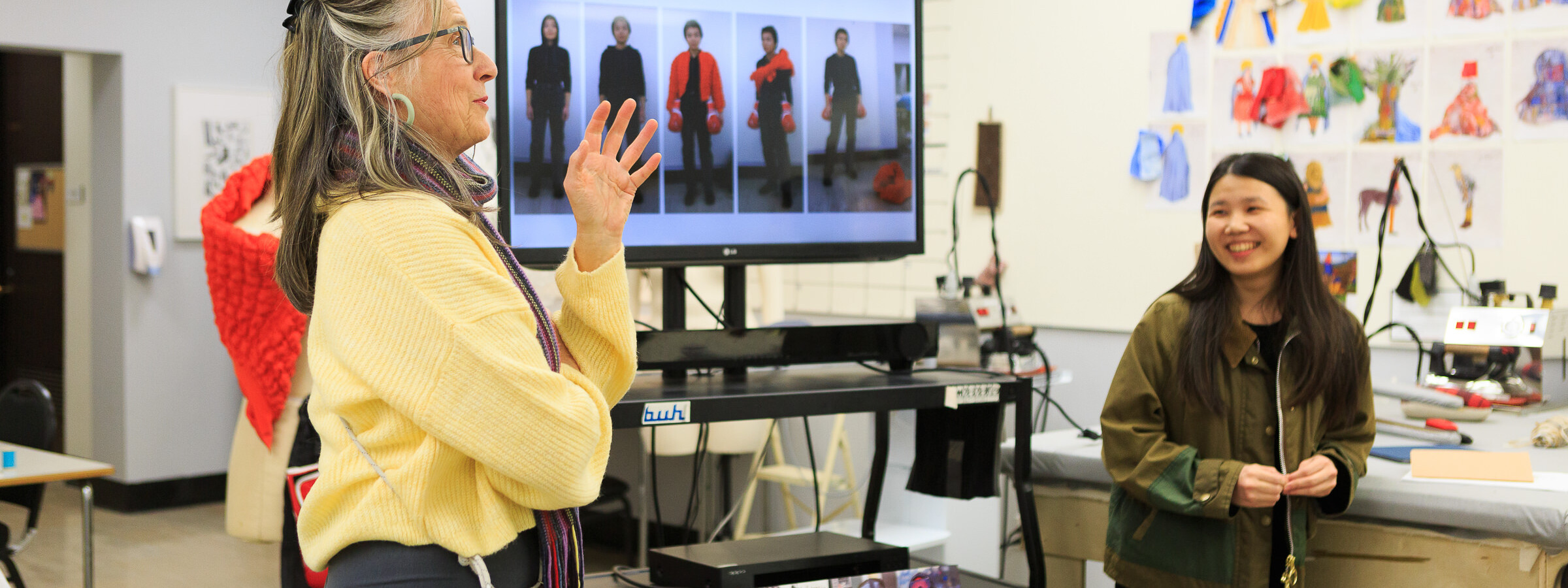 Two women standing in a workspace surround by magazines and illustrations as they review a monitor screen featuring fashion styles