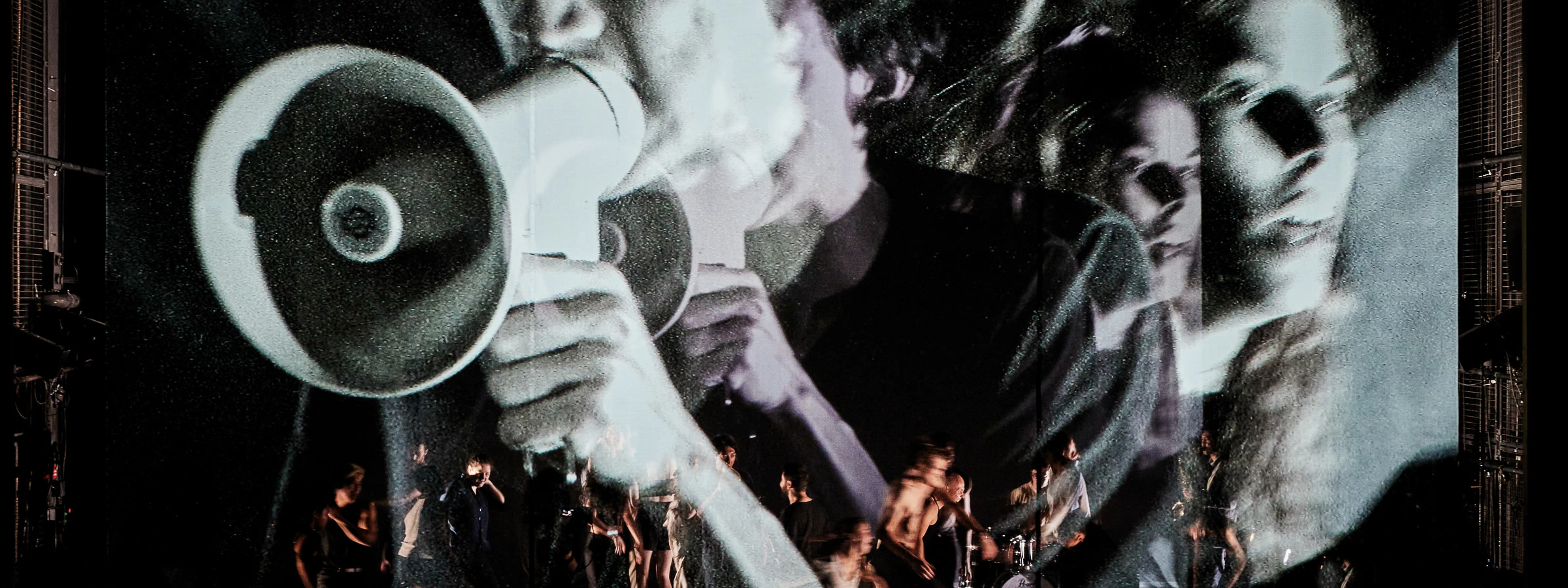 Image of a man holding a megaphone projected in black and white on a large screen as people dance in motion underneath