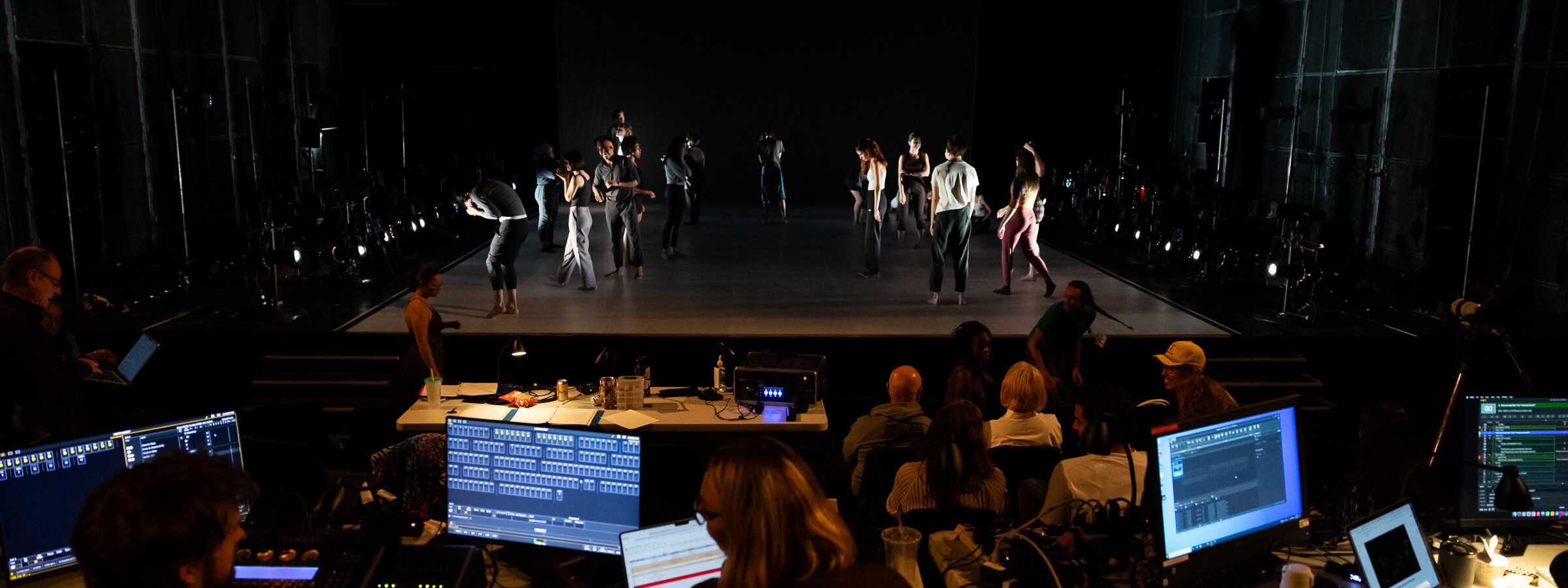 A mostly dark stage filled with performers. In front of it, people work at tables filled with production technology. The words “Augustine Machine” are projected on a screen behind the stage.