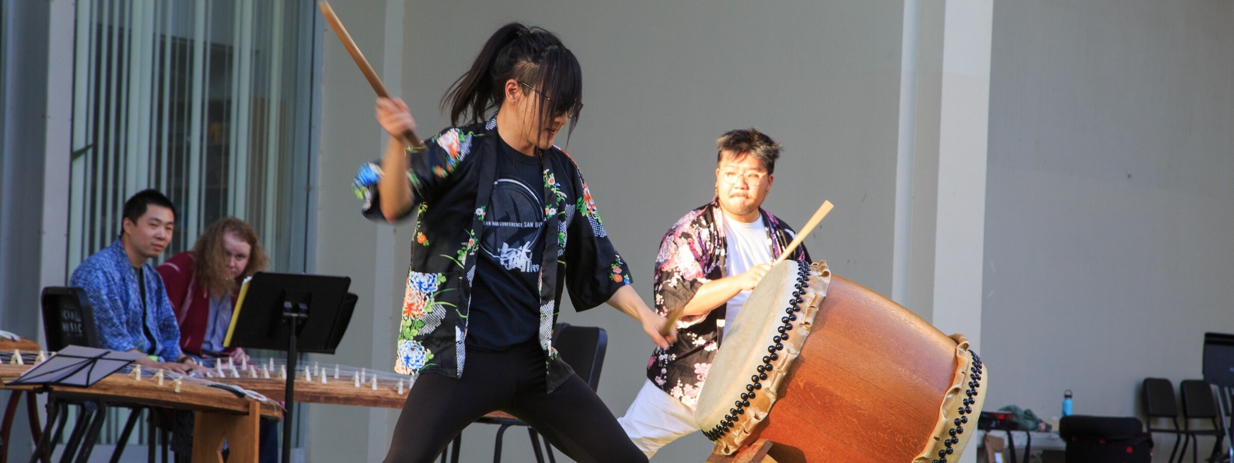 A person aggressively beats a large drum while another mirrors their pose in the background. They are outdoors and two other people sit at a stringed instrument in the background.