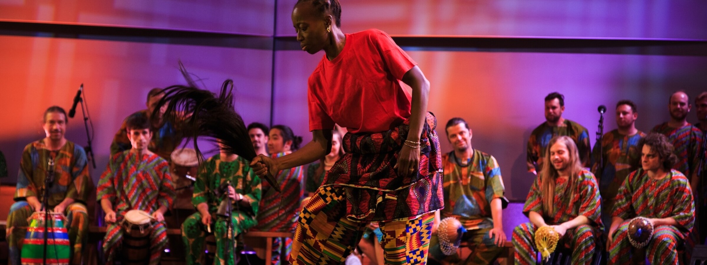 A woman in a red t-shirt and brightly colored African-patterned pants dances on a stage. A row of seated people, also dressed in bright patterned clothing, play percussion instruments behind her.