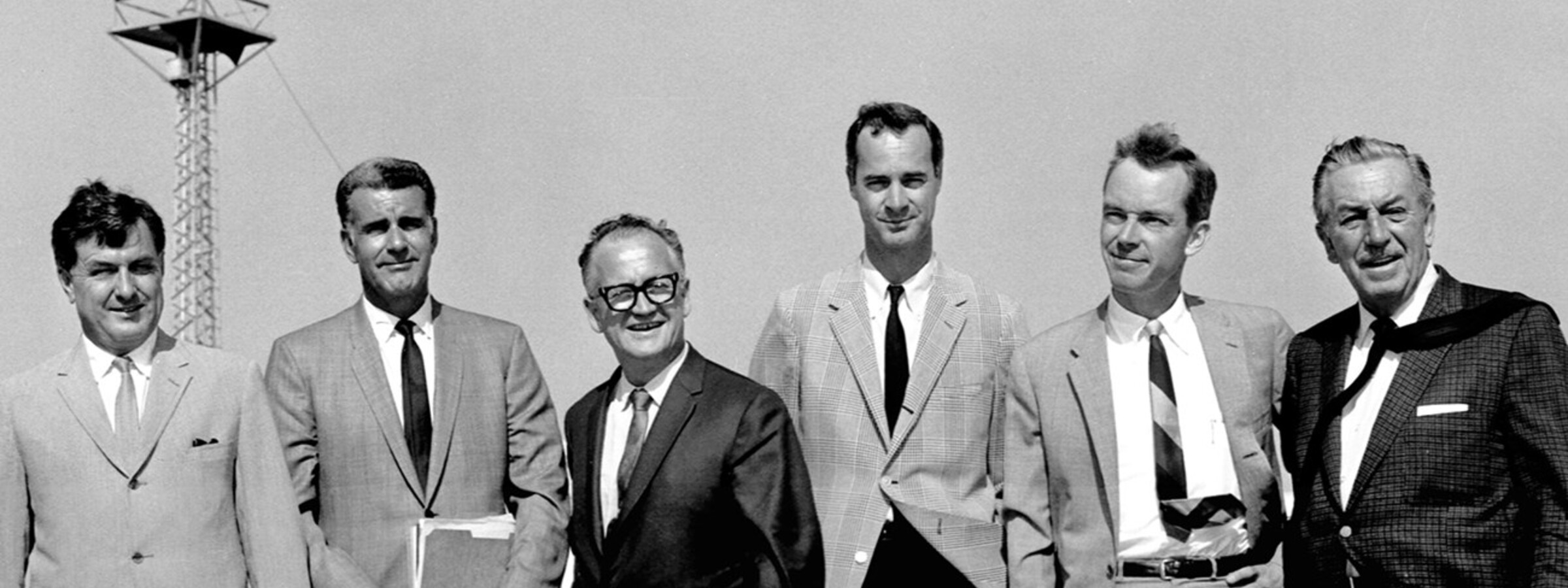 Six men in suits standing outdoors in front of a car and a tower.