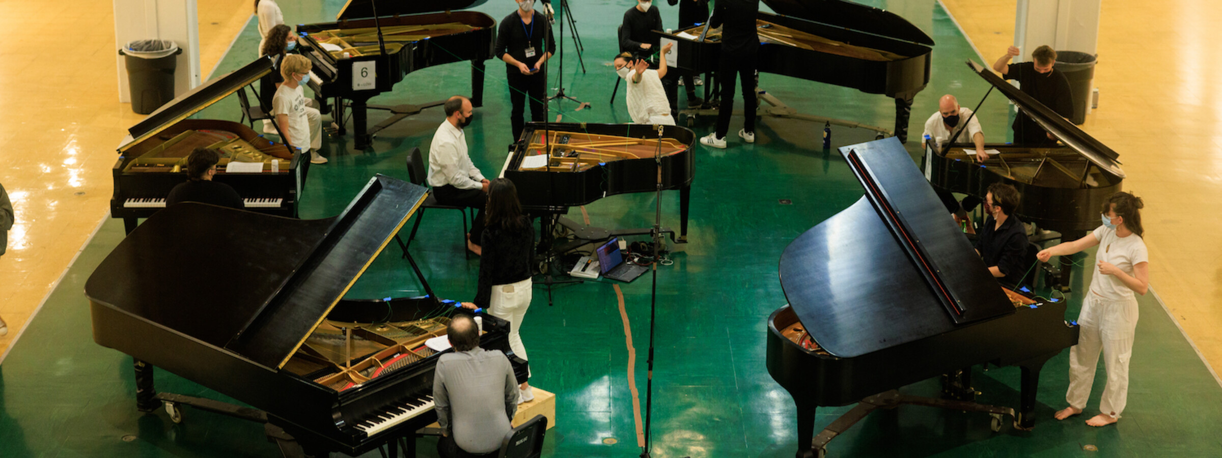 Seven pianos in a circle, each with players and dancers moving between them. 