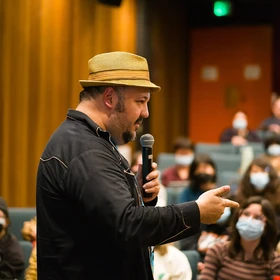 Jorge R. Gutiérrez, wearing a straw hat, speaks into a microphone to students in a small auditorium 