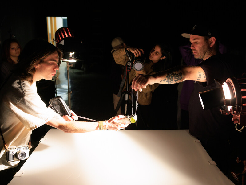 A group of four people in a dark room point photo lights and cameras at an object they’re holding above a dramatically lit white table. 