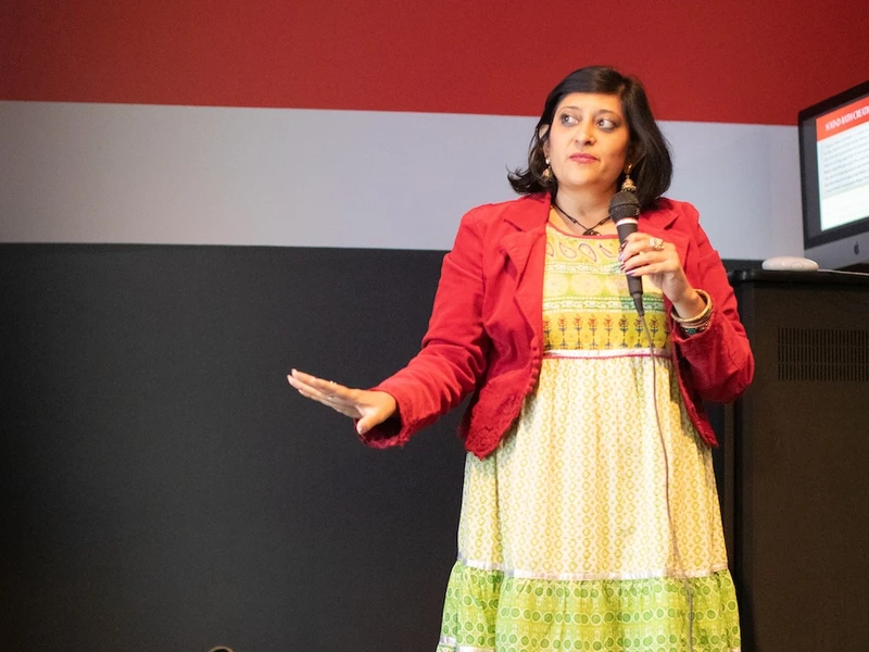 Woman in yellow dress and red jacket speaks with a microphone in her left hand 