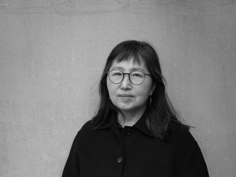 photo portrait of Don Mee Choi; 3/4 full body portrait, black and white image, of an Asian lady in all black and glasses