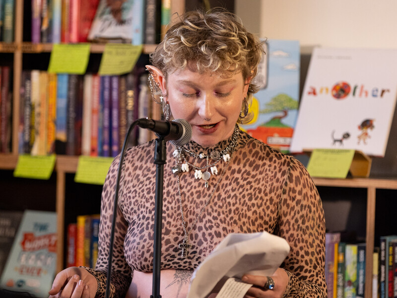 Person with short curly hair and multiple earrings, wearing a leopard print top and layered necklaces, reading from a paper while speaking into a microphone. They are standing in front of a bookshelf filled with books, some with colorful covers.