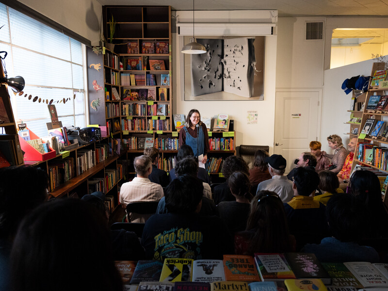 A person at a microphone addresses a small group of people seated in a cozy room lined with bookshelves. 