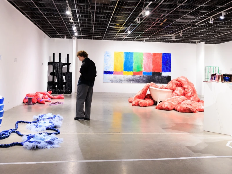 A person dressed in dark clothing looks at an art exhibition of colorful abstract sculptures and a large drawing
