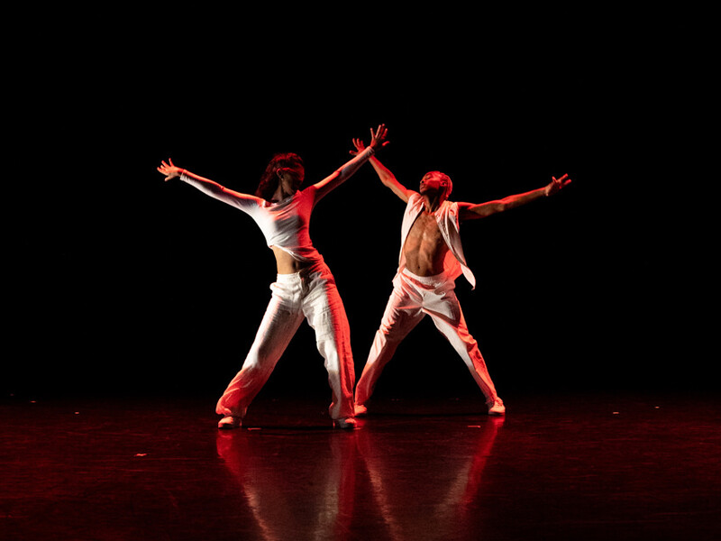 Two dancers stand on a dark stage, turned slightly toward each other, with their arms and legs spread out. 