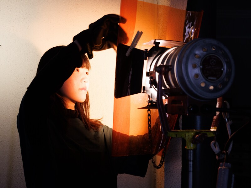 A college student adjusts a cinematic light, which shines on her face and creates an orange glow around her