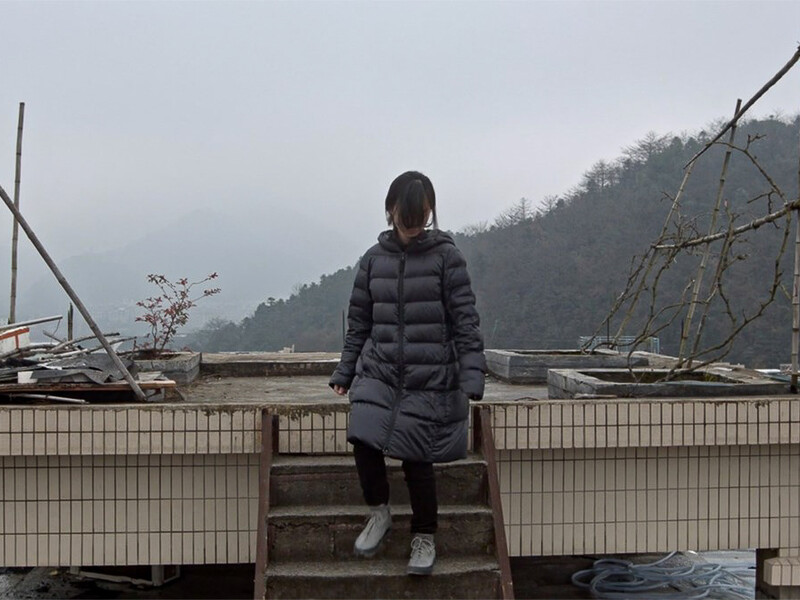 A person wearing a parka sits on a platform on a cold foggy day with mountains in the background