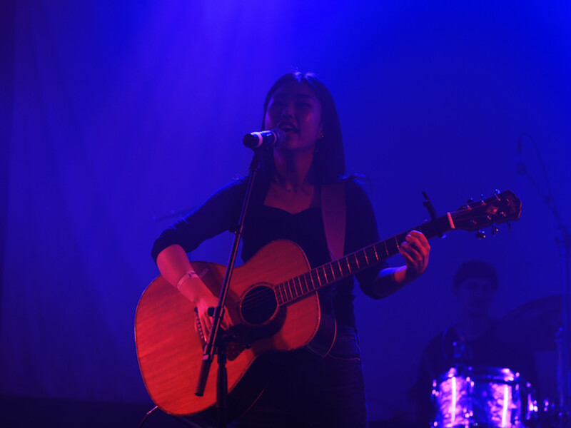 A woman plays an acoustic guitar on a blue-lit stage in a nightclub