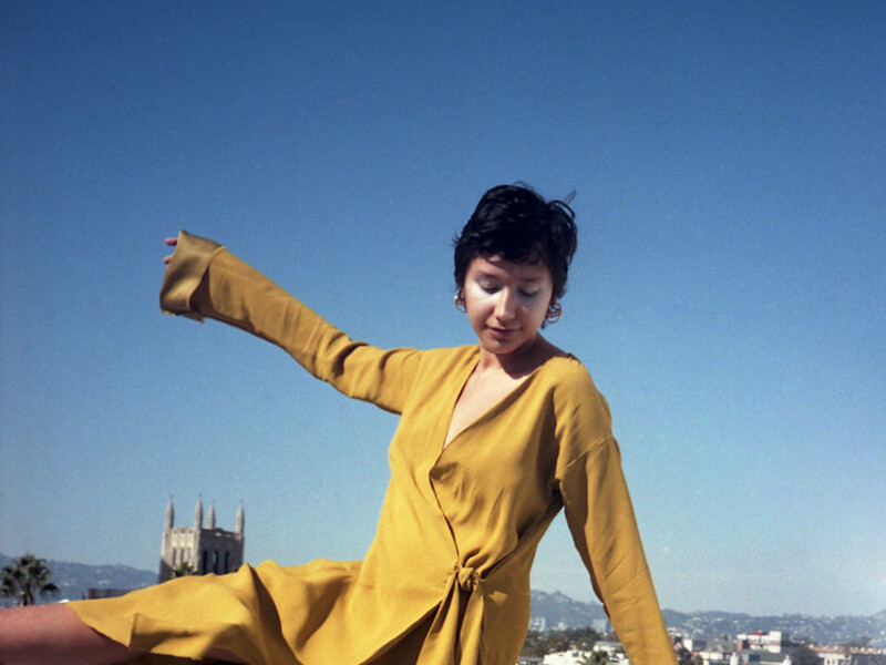 headshot of Nikki Ochoa, a woman in a yellow dress dances on a roof overlooking a city.