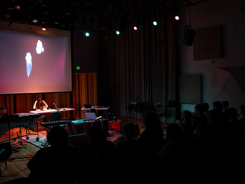 A lone person sits at a table on a stage surrounded by musical instruments and technology, a screen projection behind her. A small audience watches.