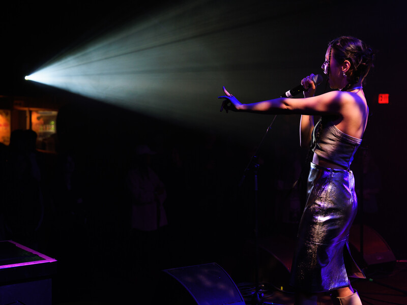 A woman sings into a microphone while holding one arm outstretched. Light from a projector streams toward her in a darkened space.