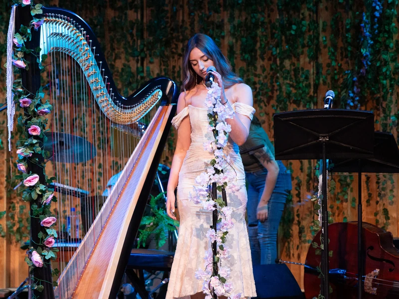 A young woman in a white dress stands at a microphone. Next to her is a large harp wrapped in flowers.