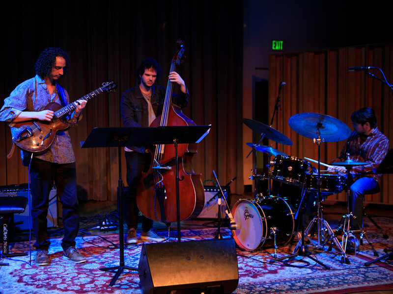 Three musicians performing on stage with a guitar, double bass, and drum kit.