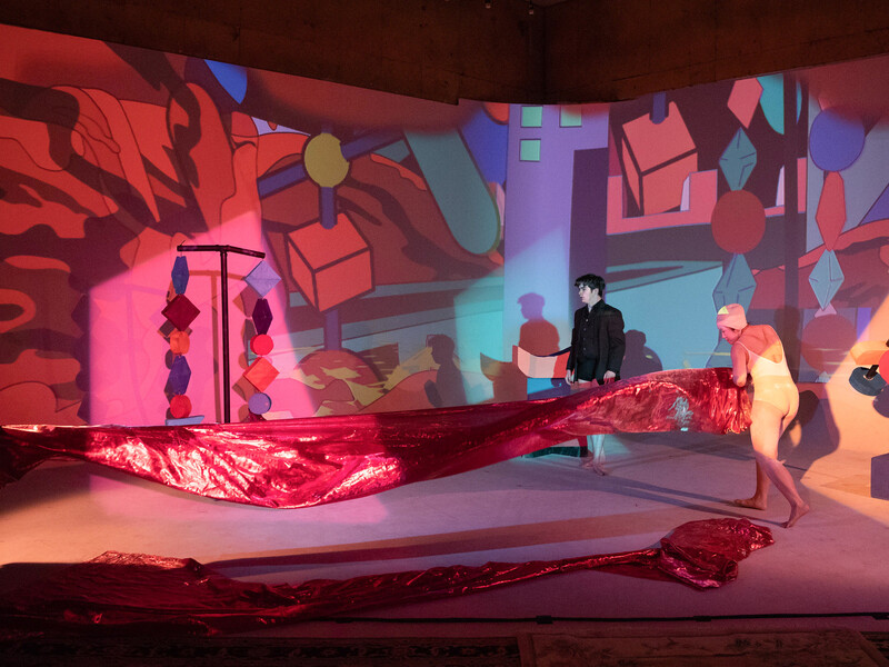 Performers stand on a brightly colored stage, holding a large banner. The stage is covered in geometric shapes. 