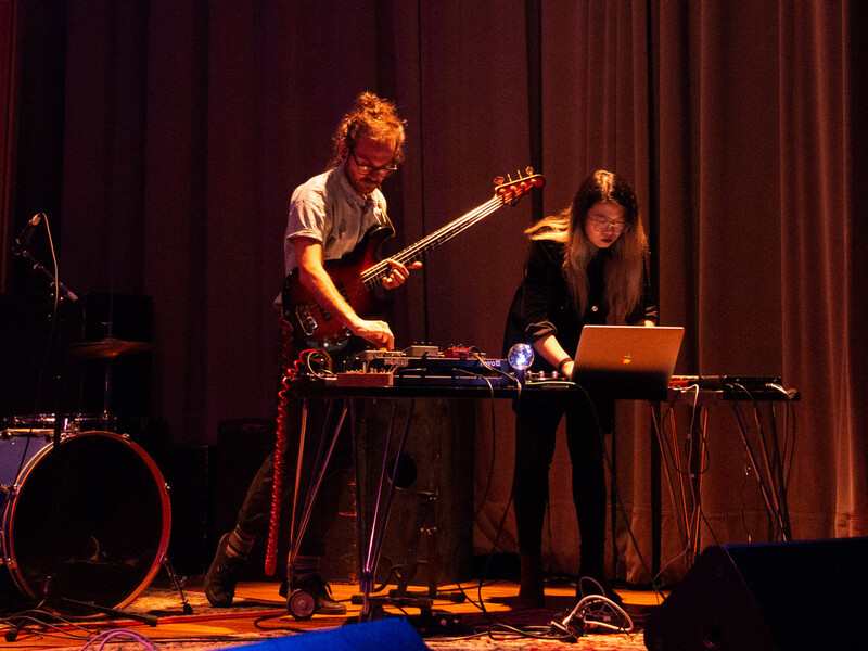 Two individuals performing on stage with musical and electronic equipment. One is playing an electric bass guitar, and the other is operating a laptop.