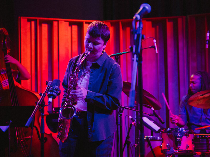 A person plays a saxophone on a small stage with moody pink and purple light. Musicians playing a standup bass and a drum set are in the background.