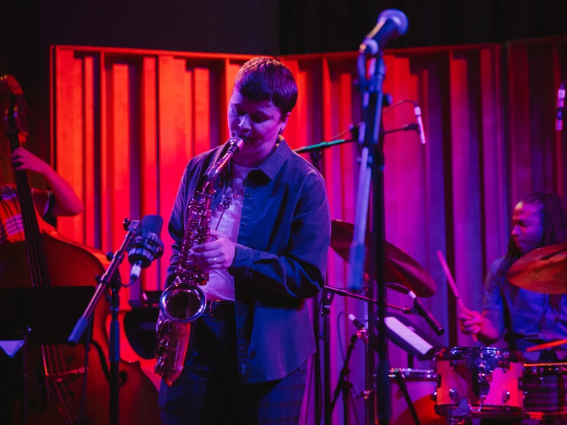 A person plays a saxophone on a small stage with moody pink and purple light. Musicians playing a standup bass and a drum set are in the background.