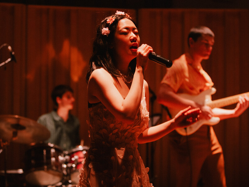 A woman in a flower crown sings intensely into a microphone, bathed in pink light. 