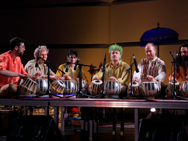 A group of tabla drum players gathers on a stage, with microphones positioned at their drums. 