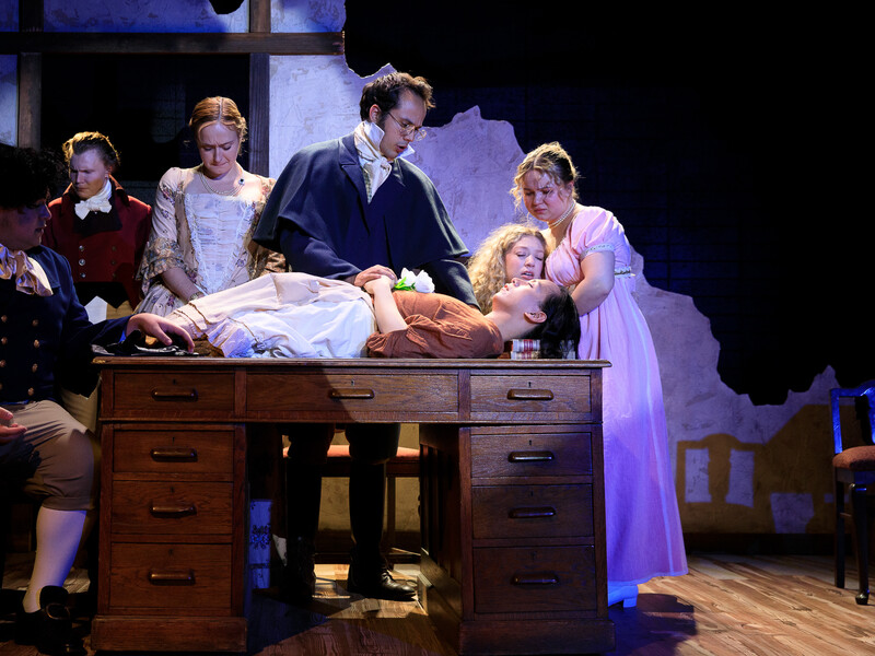 Play on a stage depicting a recently deceased person lying on a wooden desk as others surround her and mourn her body. All are dressed in colonial-era costume.