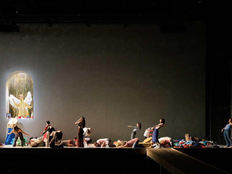 Performers on a large stage in various poses facing toward a small nook featuring a sculpture of an angel