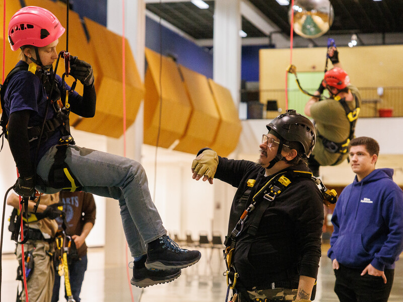 Person wearing a hard hat and strapped into a harness hangs in the air in an indoor theatrical space as another person, standing on the floor, directs them. 