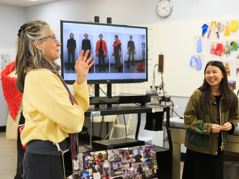 Two women standing in a workspace surround by magazines and illustrations as they review a monitor screen featuring fashion styles