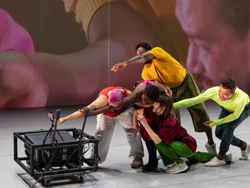 Group of people on stage posing in front of a camera as their image is projected on the wall behind them