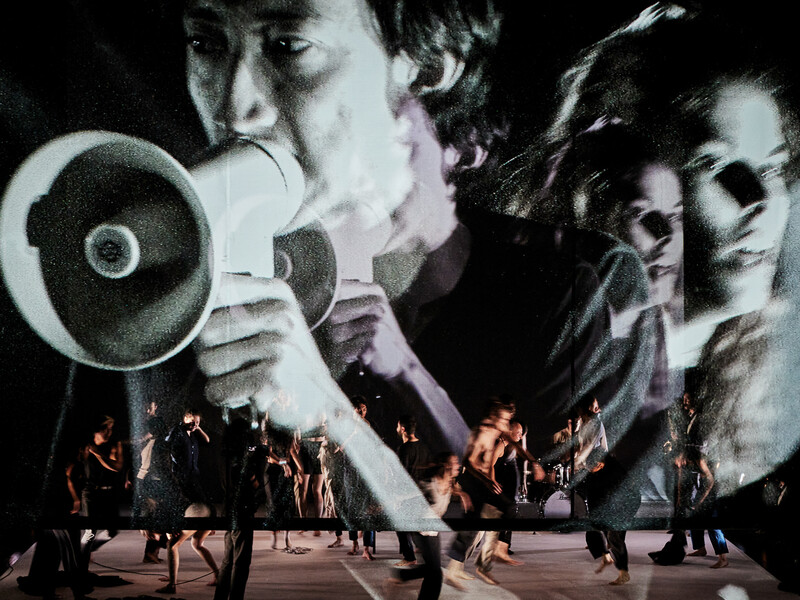 Image of a man holding a megaphone projected in black and white on a large screen as people dance in motion underneath