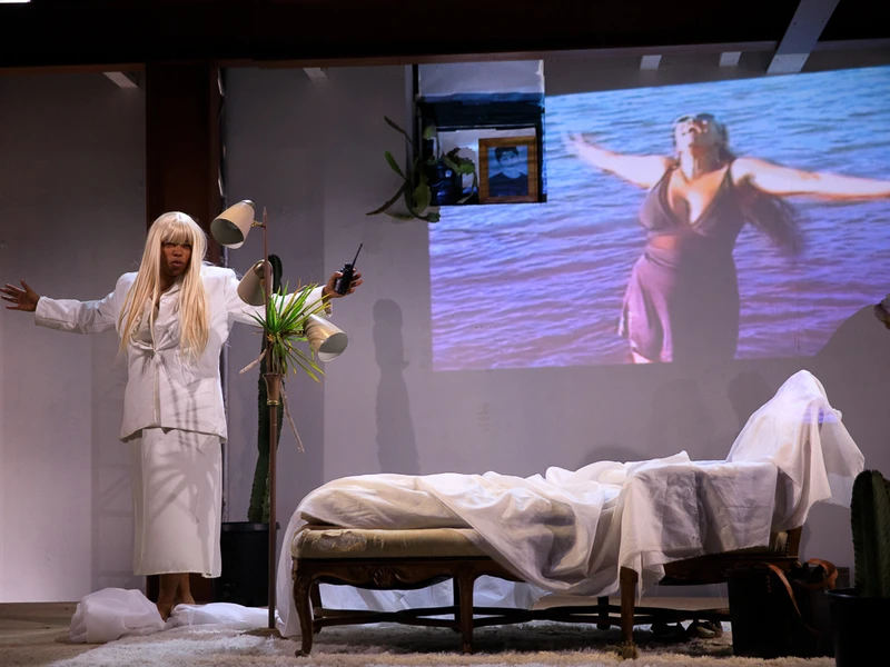 A woman wearing all white with arms outstretched in a makeshift bedrooms with another image of a woman projected on the wall 