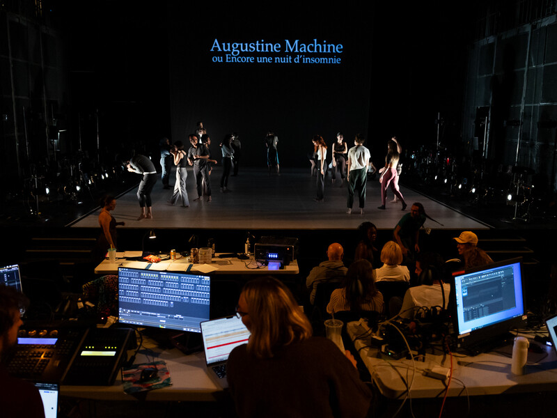 A mostly dark stage filled with performers. In front of it, people work at tables filled with production technology. The words “Augustine Machine” are projected on a screen behind the stage.