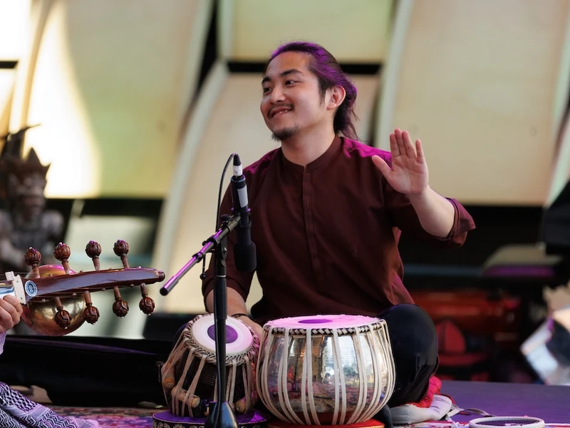 A person sits cross-legged on a patterned carpet on a stage, playing tabla and smiling