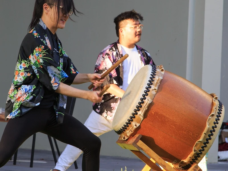 A person aggressively beats a large drum while another mirrors their pose in the background.