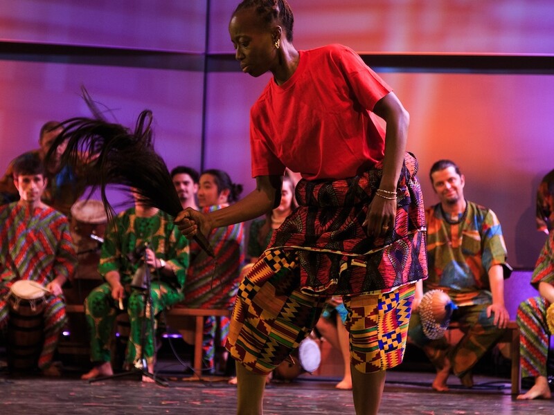 A dancer in a red shirt and colorful shorts performs with a dark prop in hand, while seated musicians in vibrant attire play in the background.