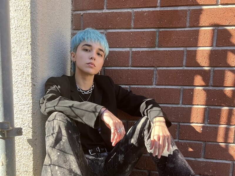 Profile photo of Wes Weisbaum, showing a person sitting in front of a brick wall, with blue hair. 