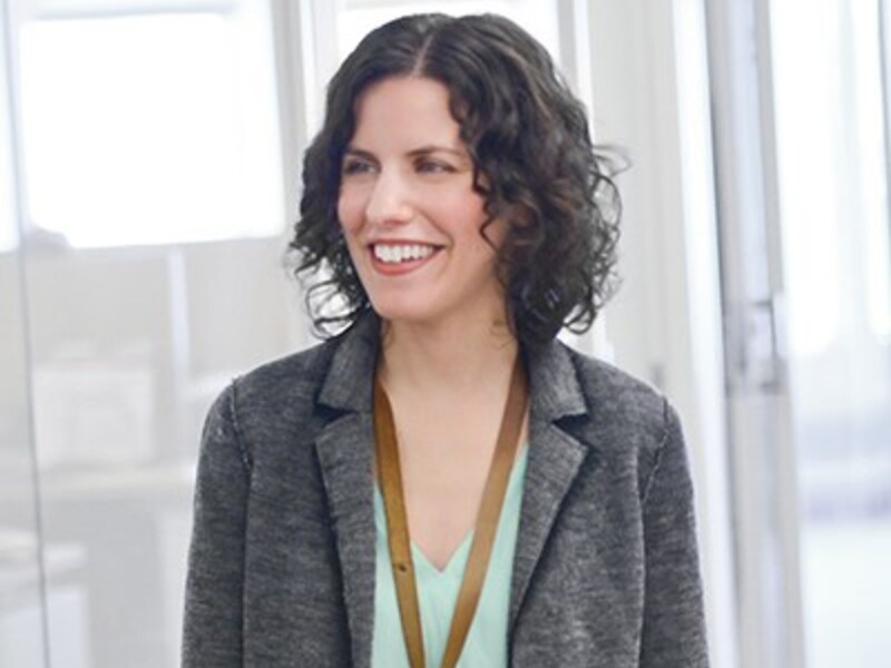 A woman with curly hair and a gray blazer smiles in an office setting.