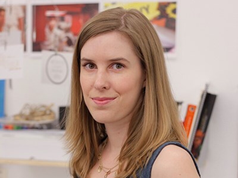 Woman with shoulder-length brown hair in a blue sleeveless top, standing indoors with blurred photos and books in the background.