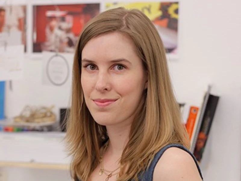 Woman with shoulder-length brown hair in a blue sleeveless top, standing indoors with blurred photos and books in the background.