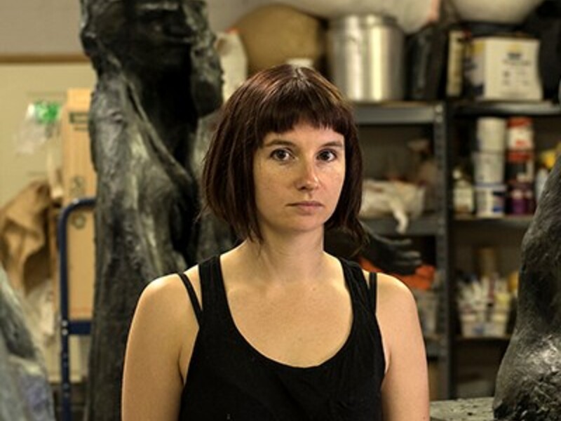 Person in an art studio with large sculptures and art supplies in the background.