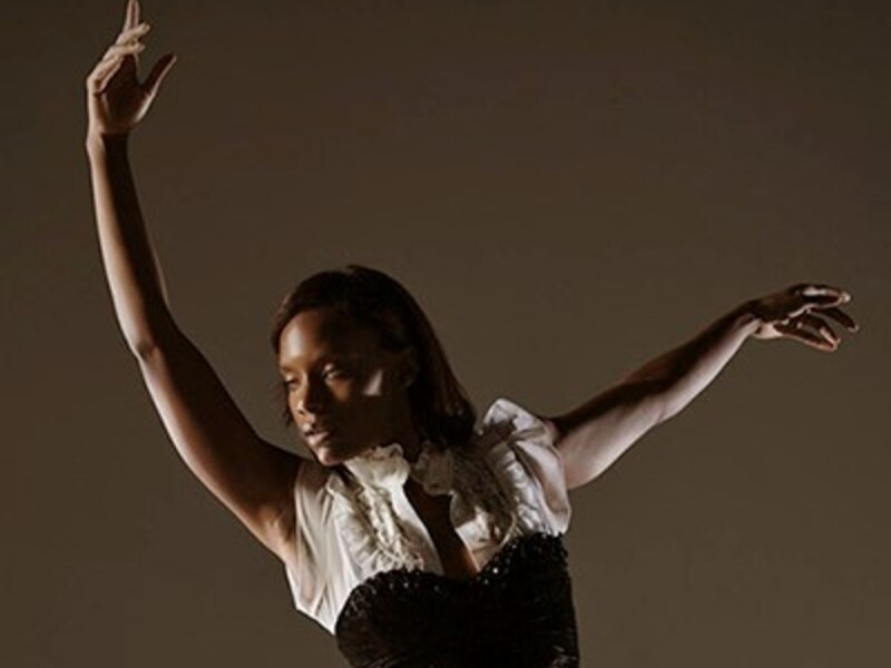 Person in a graceful dance pose with one arm raised, wearing a white top and black corset, against a brown background.