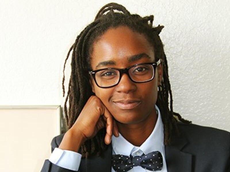 Person with dreadlocks and glasses wearing a navy suit and polka dot bow tie.