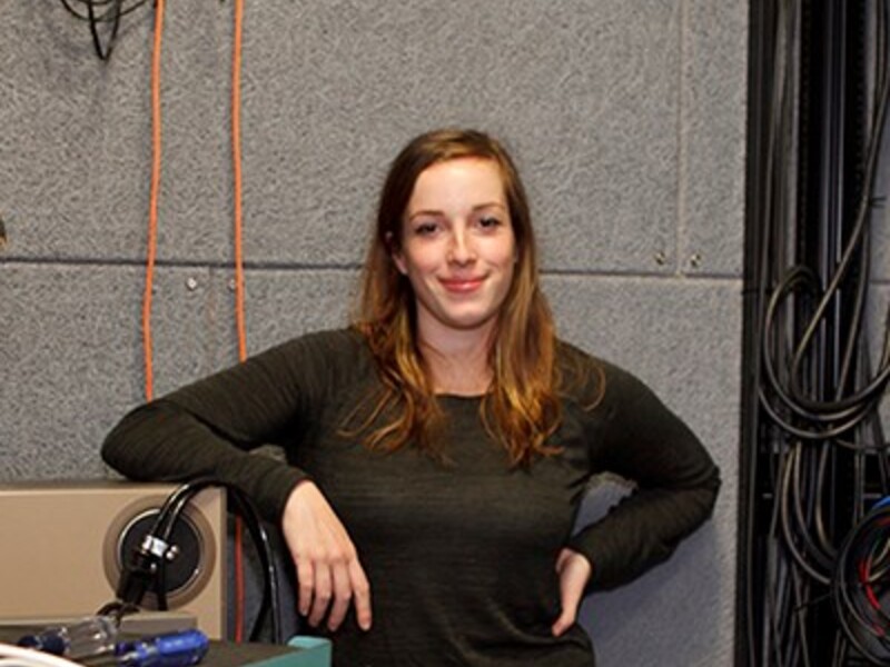 Woman standing in a workshop with technical equipment and cables in the background.