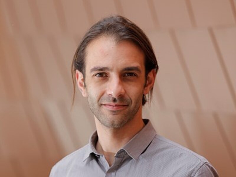 Person with long dark hair wearing a gray shirt against a light brown panel background.