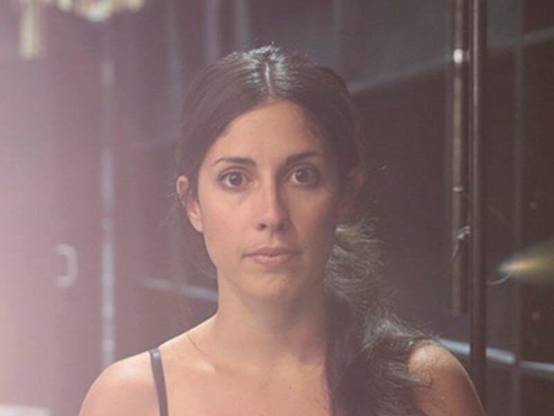 Woman in a dark setting with soft lighting, wearing a black sleeveless top.